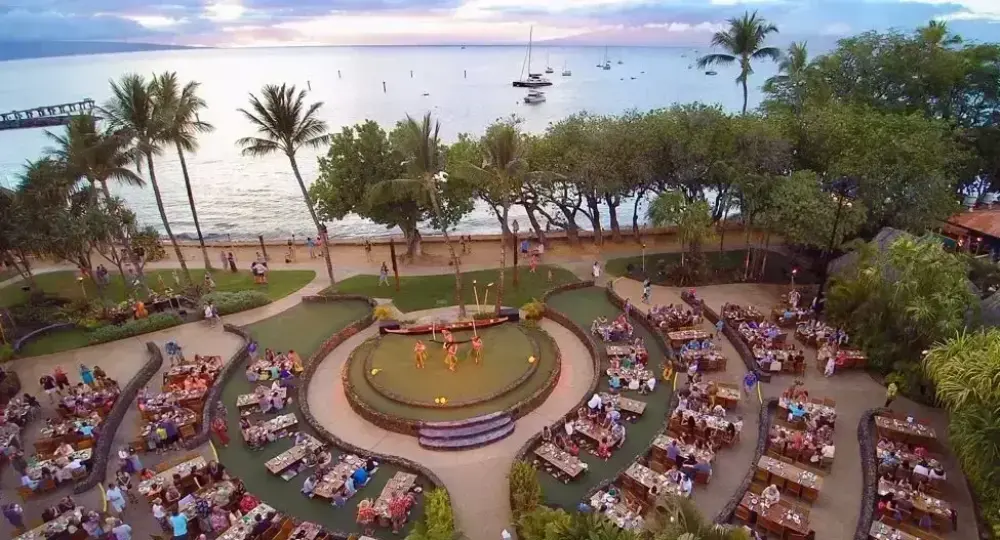 Outdoor luau stage with performers surrounded by dining guests near ocean at sunset