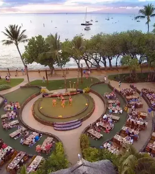 Outdoor luau stage with performers surrounded by dining guests near ocean at sunset