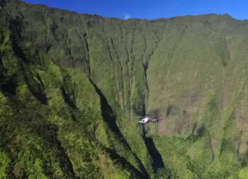 Helicopter flying near lush green steep mountains under clear blue sky