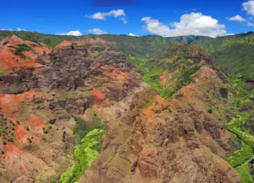 Waimea Canyon with red and brown rocky cliffs and lush green vegetation under a bright blue sky with scattered clouds