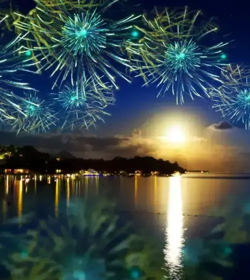 Nighttime fireworks over a calm coastal scene reflecting lights on water