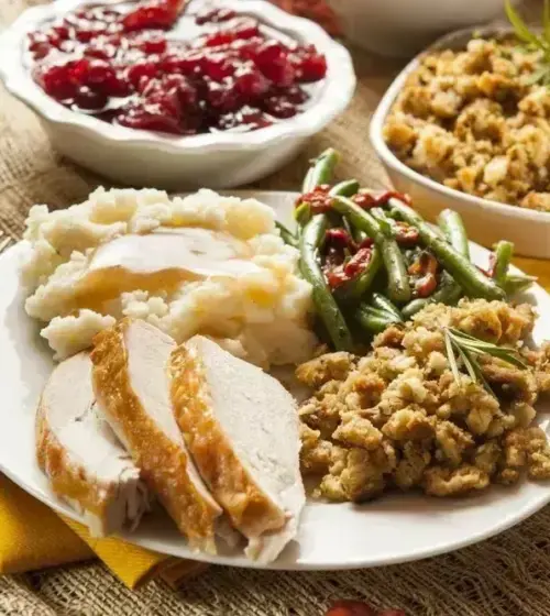 Plate with sliced roasted turkey, mashed potatoes with gravy, green beans with red peppers, stuffing, cranberry sauce, and corn