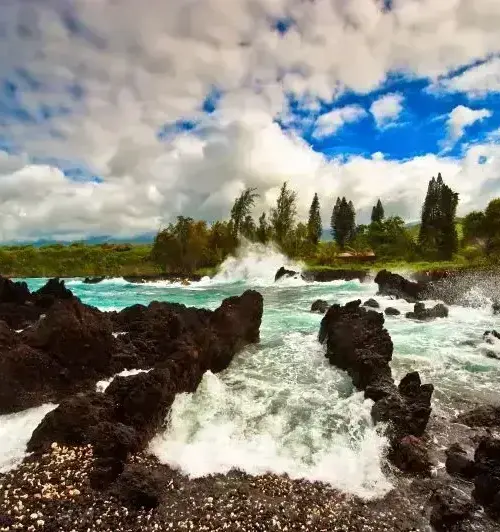 Volcanic rock formations along the Hawaiian shoreline with ocean waves crashing and a cloudy blue sky
