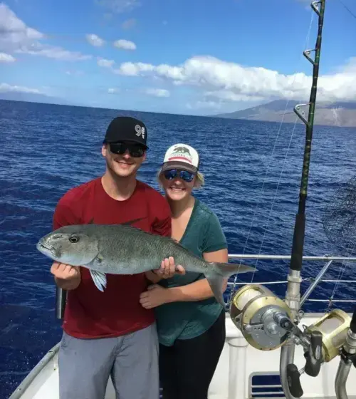 Smiling couple holding a large fish on a boat with ocean and mountains in background