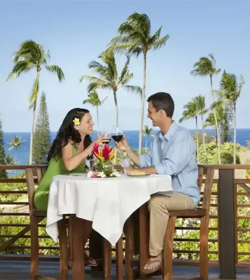 Couple clinking wine glasses at a table on a balcony overlooking ocean and palm trees