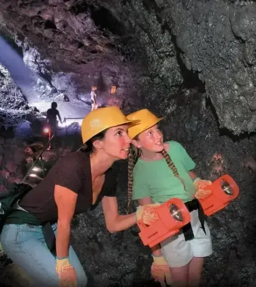 Two tourists with helmets and gloves exploring a lava tube cave holding orange flashlights