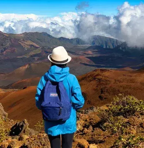 hiker in blue jacket and white hat looking over volcanic landscape