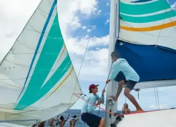 Two crew members adjusting colorful sails on a catamaran in open ocean under partly cloudy sky