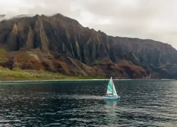 Sailboat with colorful sails on ocean near rugged cliffs under cloudy sky