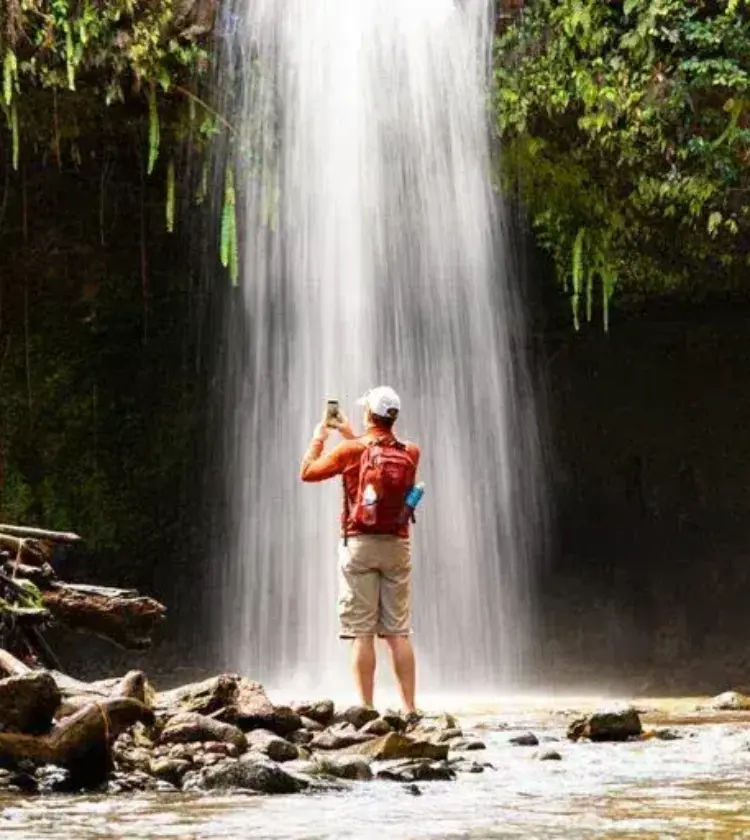 Person in orange jacket photographing waterfall in lush forest