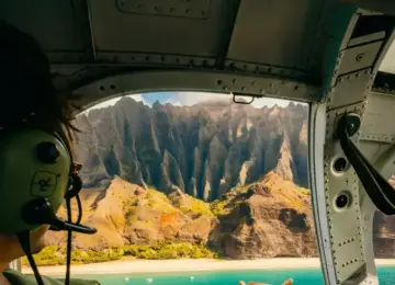 Person wearing headset inside helicopter looking out at Na Pali Coast with mountain cliffs and turquoise ocean