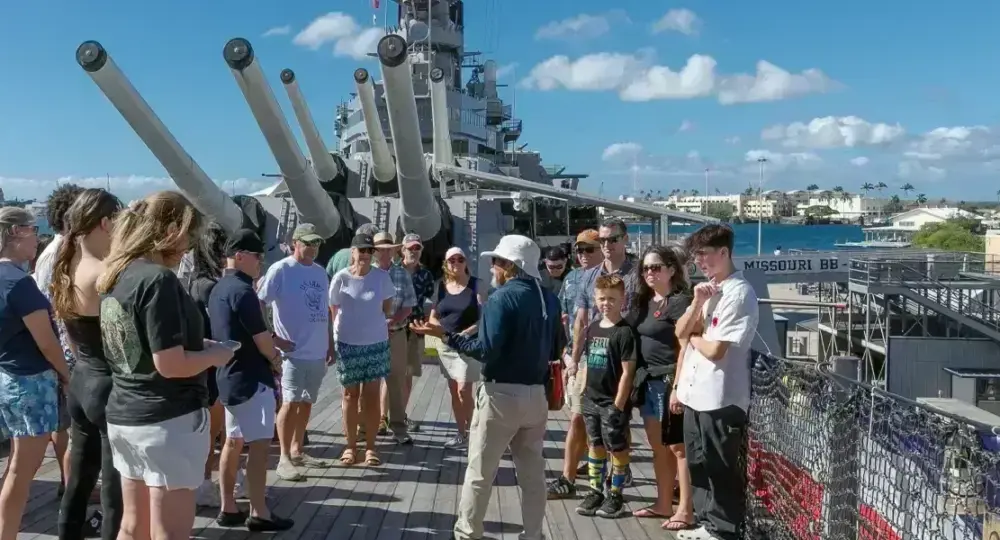 Group of tourists listening to guide on deck of a battleship with large guns and waterfront in background