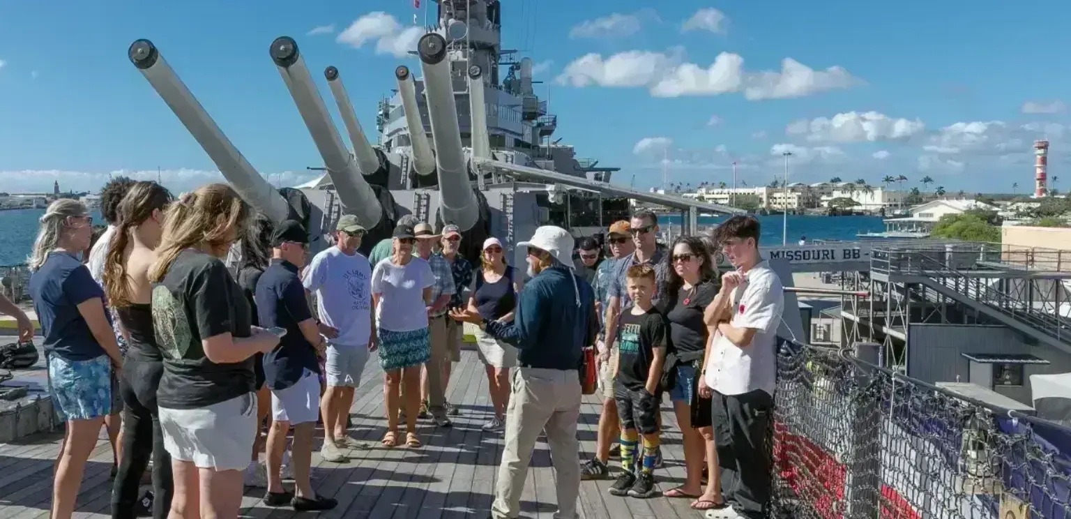 Group of tourists listening to guide on deck of a battleship with large guns and waterfront in background