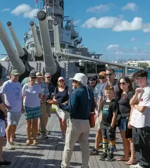Group of tourists listening to guide on deck of a battleship with large guns and waterfront in background