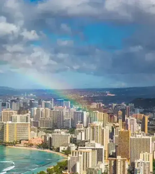 Honolulu skyline along turquoise ocean with rainbow and cloudy sky