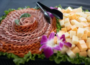 charcuterie platter with sliced deli meats shaped in a rose, cubes of yellow and white cheese, green lettuce, and purple orchid flowers