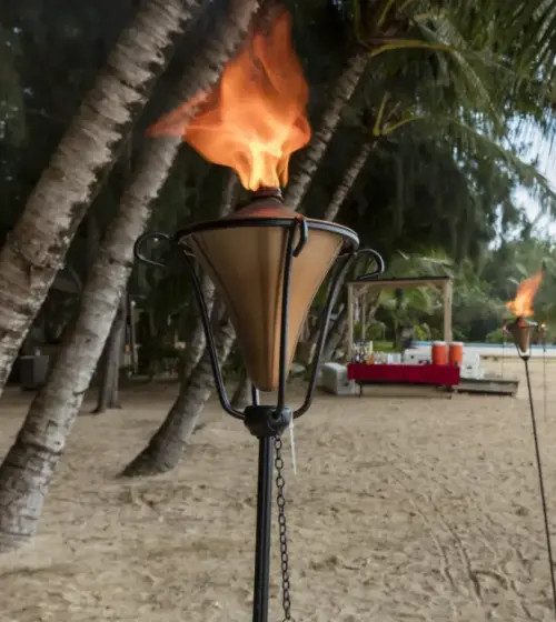Tiki torch glowing on a sandy Hawaiian beach with palm trees and ocean view with distant small island