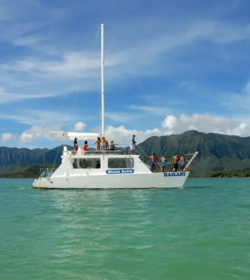 white sailboat with people on deck floating on turquoise water with forested mountains and partly cloudy sky