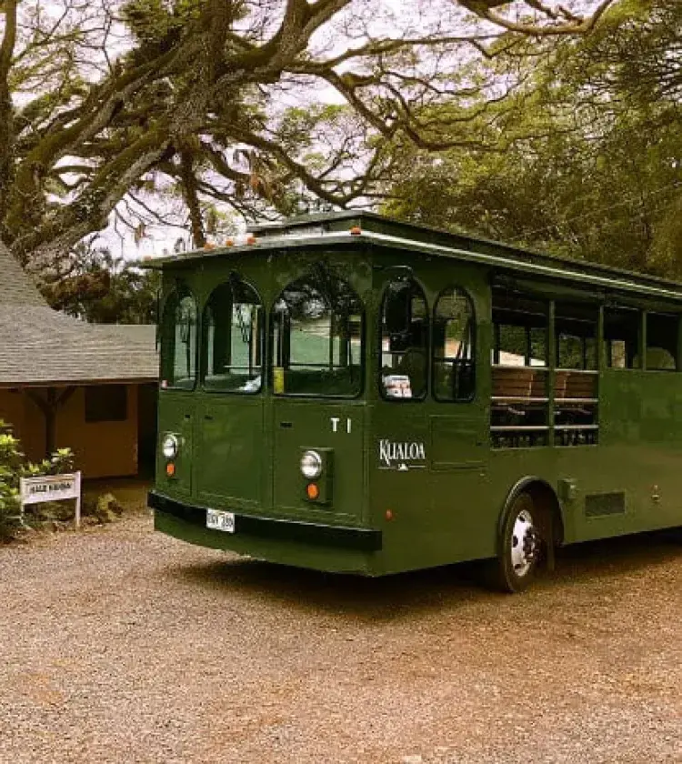 Green Kualoa Ranch tour trolley parked outside rustic building under large trees