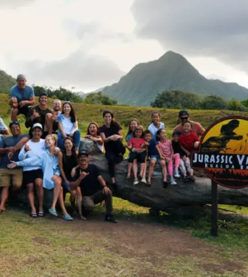 Large group posing on and around a log with Jurassic Valley Makua Ranch sign and mountain in background