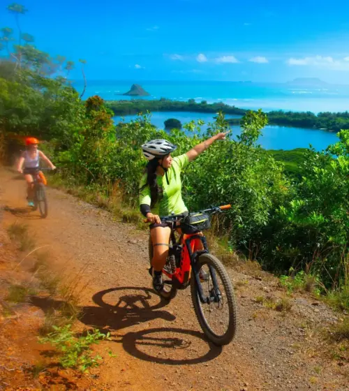 woman wearing helmet riding mountain bike on dirt trail overlooking blue ocean and green island