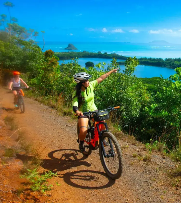 woman wearing helmet riding mountain bike on dirt trail overlooking blue ocean and green island