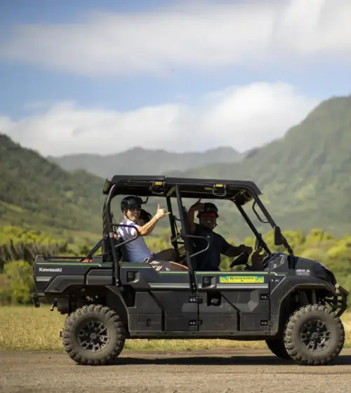 Two people riding a black Kawasaki ATV with a mountain and Moai statue in the background under cloudy sky