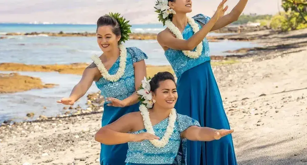 Three Hawaiian women in blue dresses performing traditional dance on beach