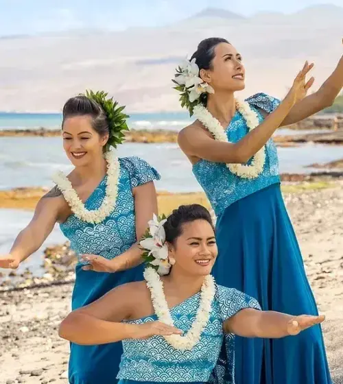 Three Hawaiian women in blue dresses performing traditional dance on beach