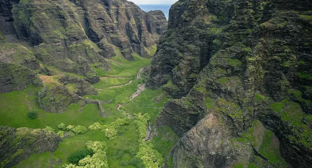 Green mountainous valley with rocky cliffs and a small stream under a partly cloudy sky