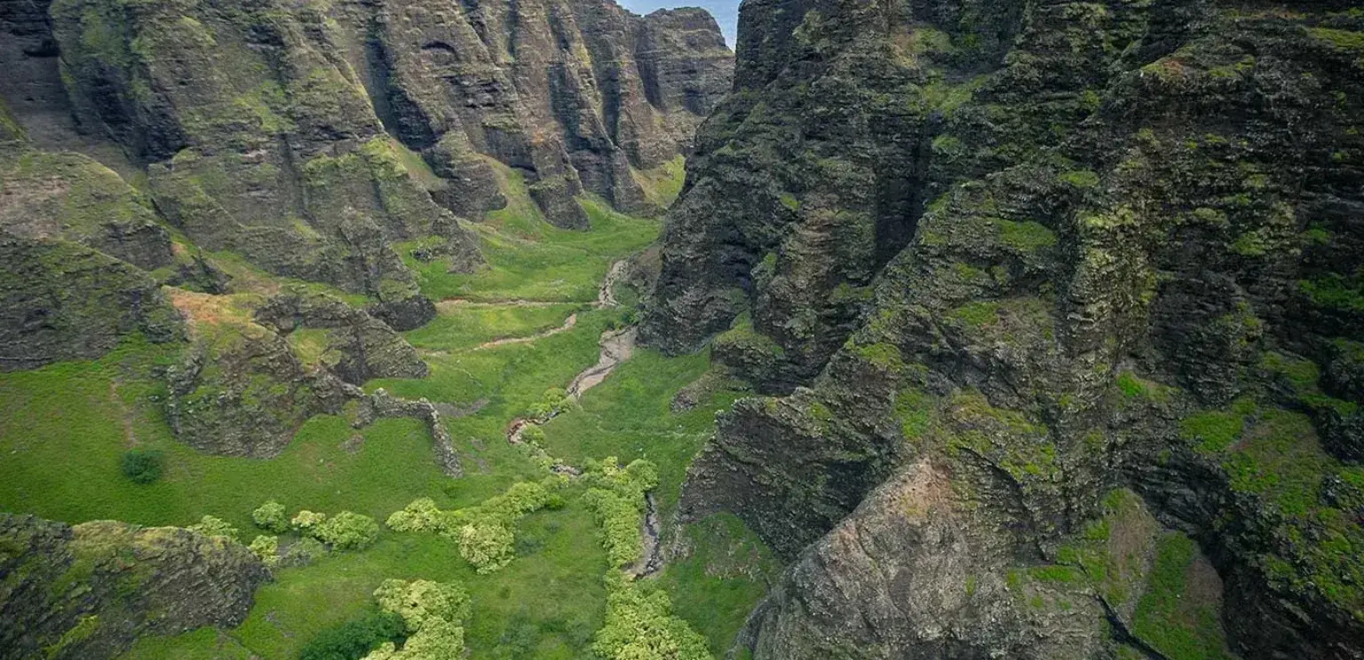 Green mountainous valley with rocky cliffs and a small stream under a partly cloudy sky
