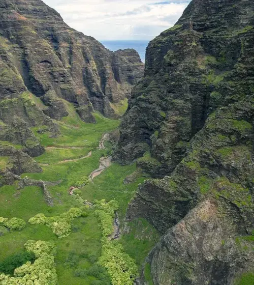 Green mountainous valley with rocky cliffs and a small stream under a partly cloudy sky