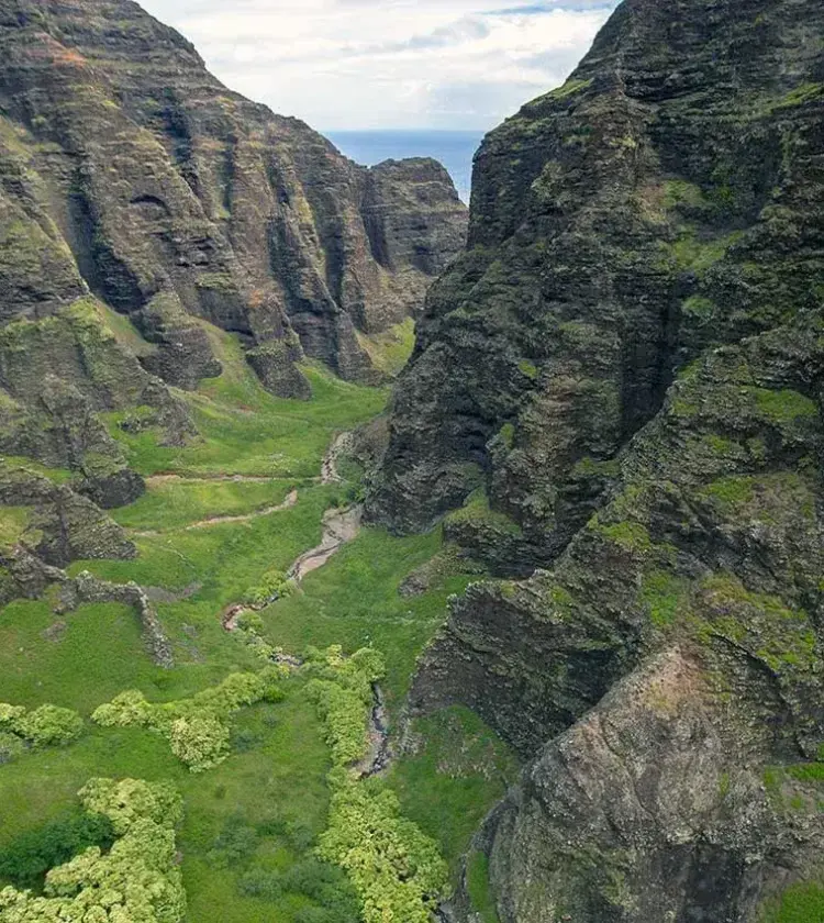 Green mountainous valley with rocky cliffs and a small stream under a partly cloudy sky