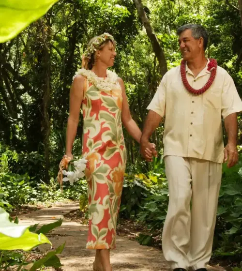 Couple holding hands walking on path through tropical forest wearing Hawaiian lei and floral dress