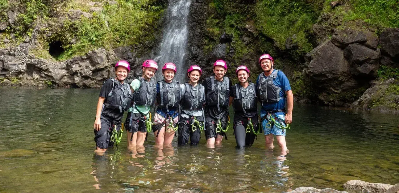 Seven adventure tourists in helmets standing in shallow water by a waterfall in a rocky, green landscape