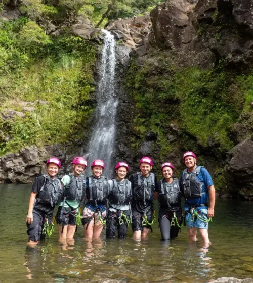 Seven adventure tourists in helmets standing in shallow water by a waterfall in a rocky, green landscape