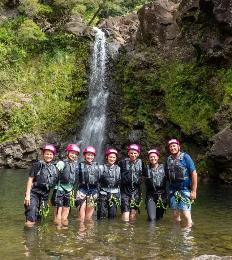 Seven adventure tourists in helmets standing in shallow water by a waterfall in a rocky, green landscape