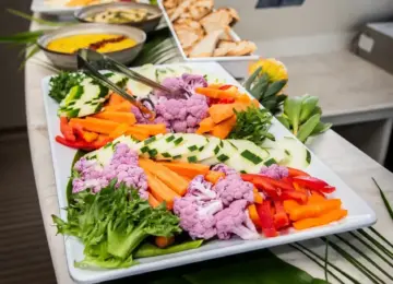 Colorful fresh vegetables including purple cauliflower, cucumbers, carrots, and peppers on a platter with bread and dips on a boat table