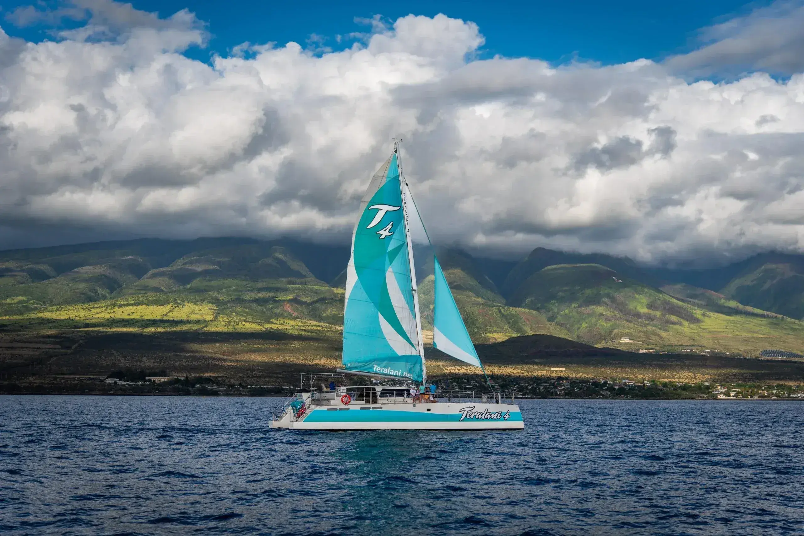turquoise and white sailboat sailing on ocean in front of lush green mountains with cloudy sky
