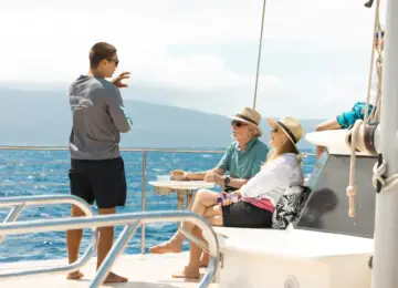 Boat tour guide talking to two seated passengers wearing hats on a yacht with ocean and mountains in background