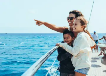 woman and two children smiling and pointing while standing on boat railing with ocean background