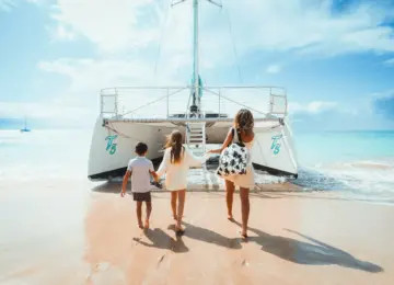 mother and two children walking barefoot on sandy beach towards large white catamaran