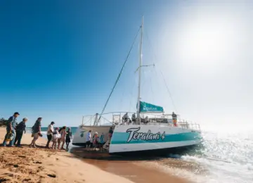 people boarding Teralani 4 catamaran on sandy beach with bright blue sky