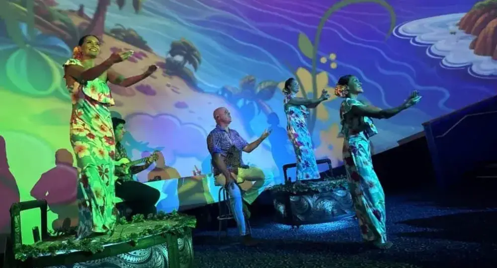 Hawaiian dancers in floral dresses with musician under colorful tropical wall projection