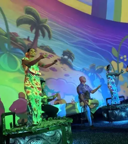 Hawaiian dancers in floral dresses with musician under colorful tropical wall projection