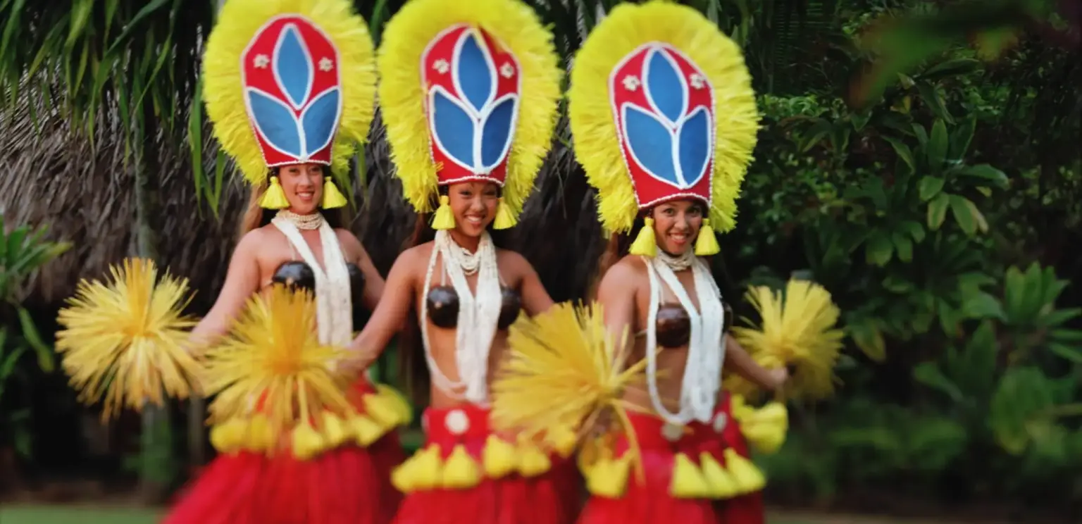 Three Hawaiian hula dancers with yellow feathered headdresses and red grass skirts dancing outdoors