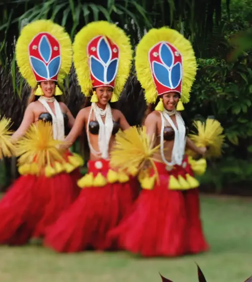 Three Hawaiian hula dancers with yellow feathered headdresses and red grass skirts dancing outdoors