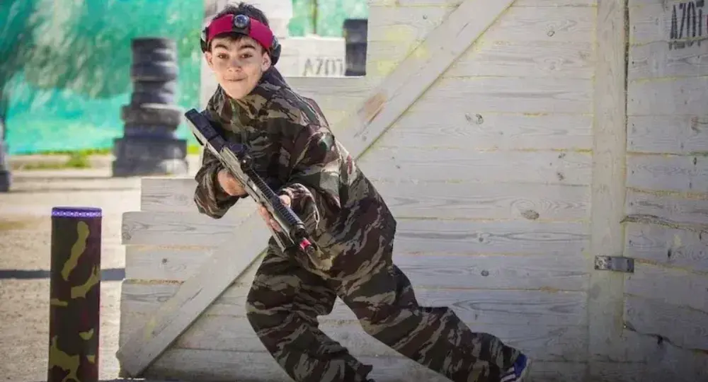 boy in camouflage playing paintball behind wooden barrier