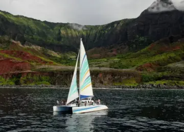 Sailboat with colorful sails carrying passengers on water near green and red forested mountains under cloudy sky