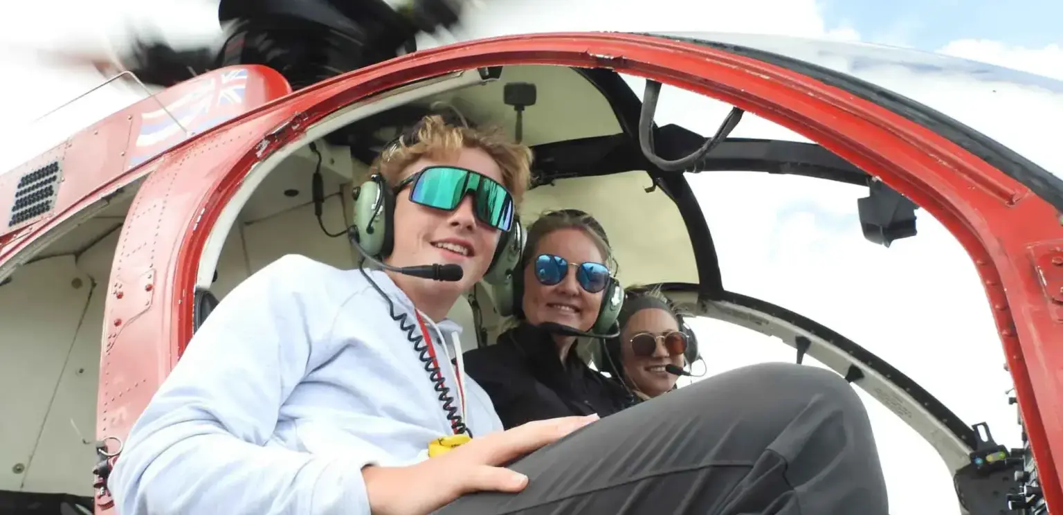 Three people wearing headsets and sunglasses sitting inside a helicopter cockpit smiling